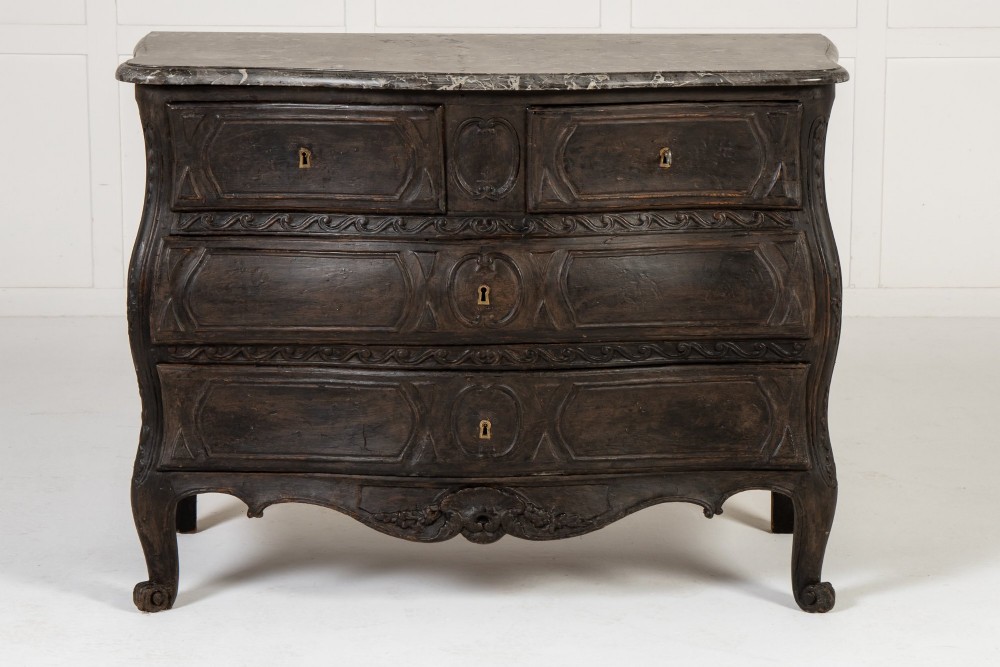 An 18th-century ebonised French bombe commode featuring three drawers, carved panels, a grey marble top, and brass keyholes.