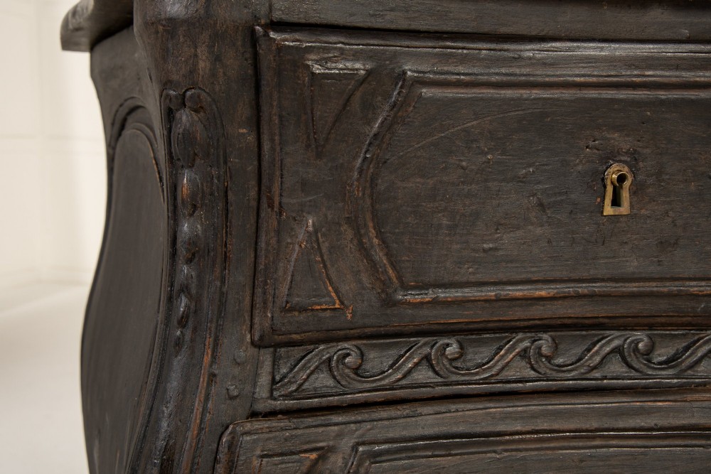 An 18th-century ebonised French bombe commode featuring three drawers, carved panels, a grey marble top, and brass keyholes. View IV