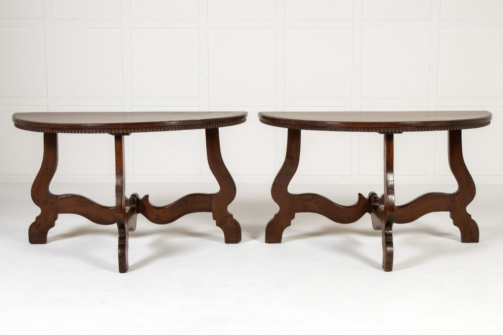 A pair of 18th-century Italian walnut demi-lune console tables with carved dentil edges, shaped legs, and central stretchers.