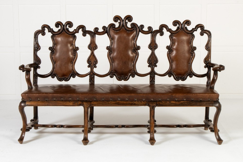 A 19th-century carved walnut settee featuring a triple chair-back design with scrollwork carving and a brown leather studded seat.