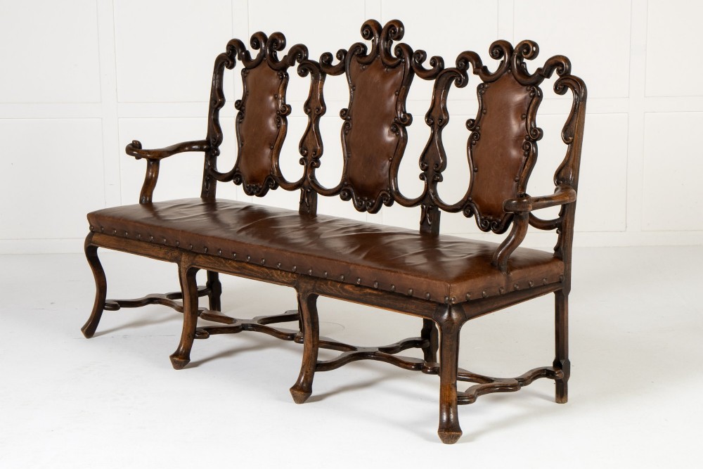 A 19th-century carved walnut settee featuring a triple chair-back design with scrollwork carving and a brown leather studded seat. View II