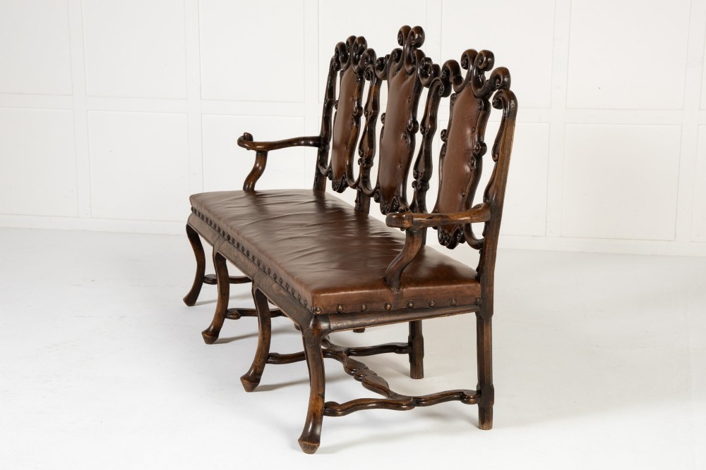 A 19th-century carved walnut settee featuring a triple chair-back design with scrollwork carving and a brown leather studded seat. View IV