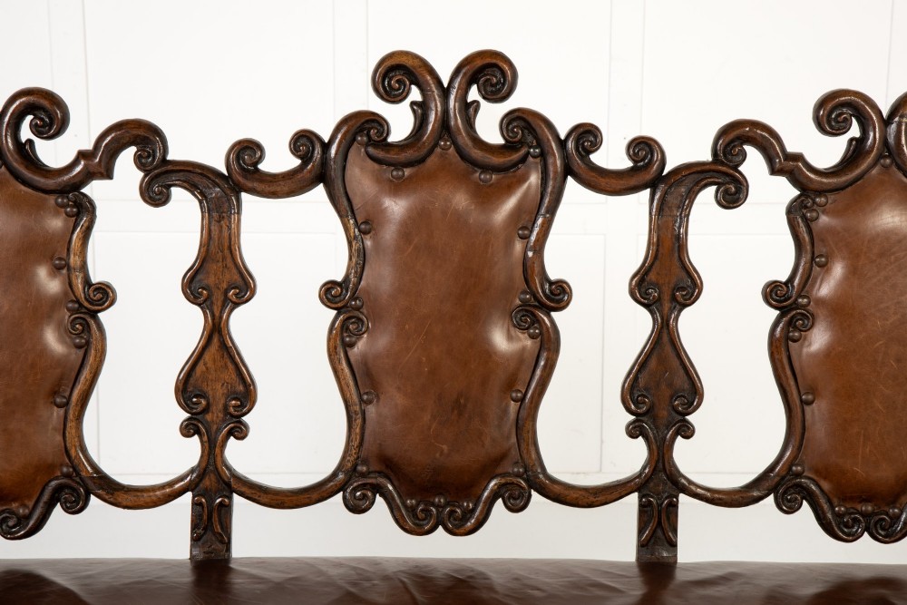 A 19th-century carved walnut settee featuring a triple chair-back design with scrollwork carving and a brown leather studded seat. View V