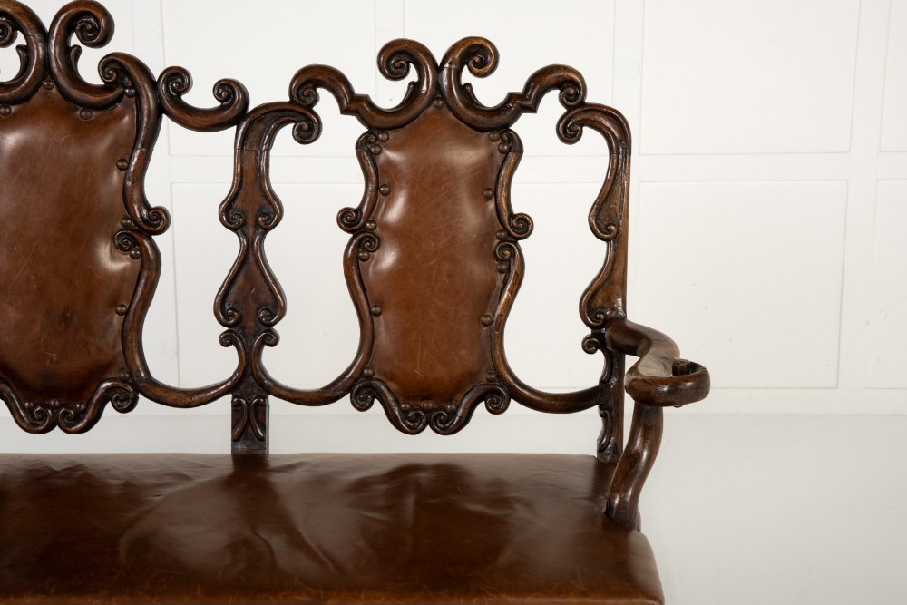 A 19th-century carved walnut settee featuring a triple chair-back design with scrollwork carving and a brown leather studded seat. View VI