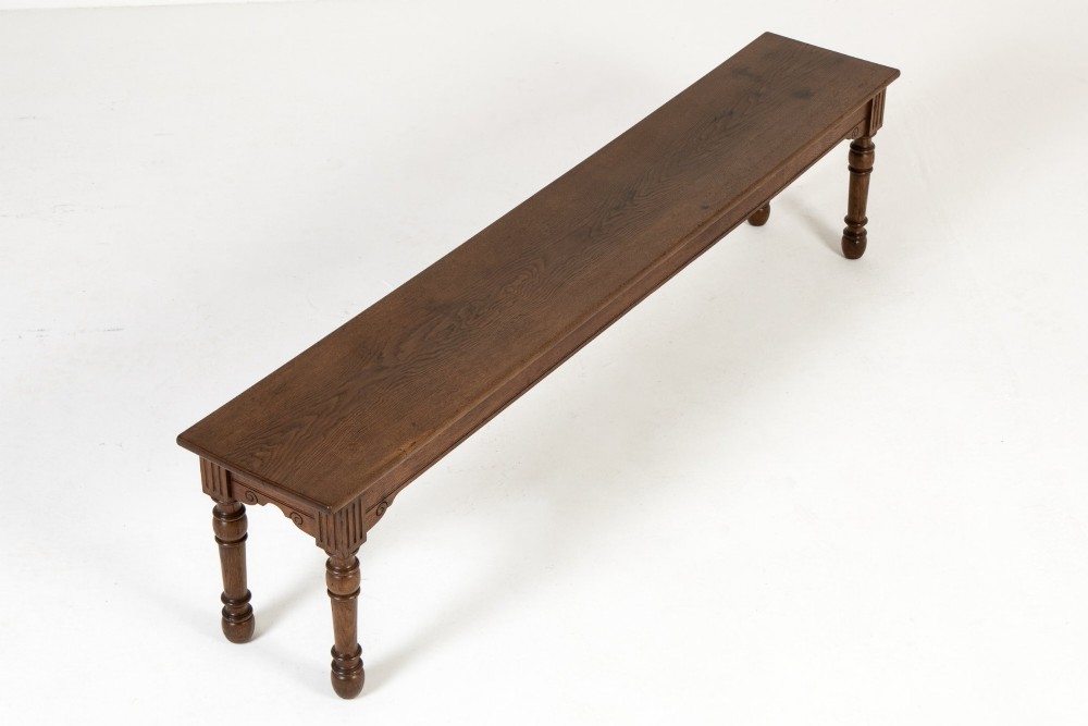 A long 19th-century dark oak bench featuring a rectangular plank seat supported by turned legs with fluted frieze details.