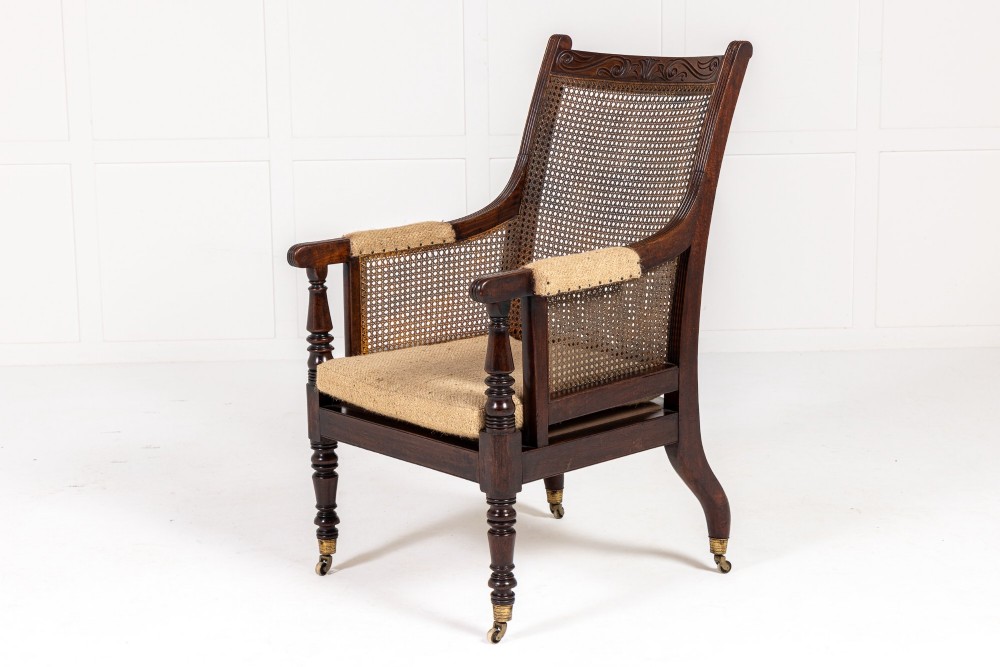 A Regency dark mahogany bergere library armchair featuring woven cane back and side panels, turned front legs on brass castors, and upholstered armrests.