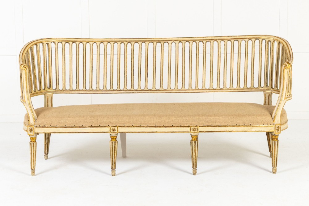 An 18th-century Italian Neoclassical settee featuring a cream-painted frame with gold leaf accents, a curved vertical spindle back, fluted turned legs, and a neutral upholstered seat.