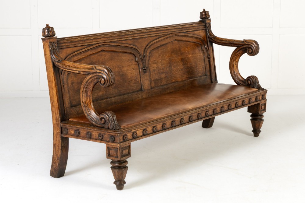 A nineteenth-century carved dark oak Gothic Revival bench featuring pointed arches on the backrest, scrolled armrests, and a fitted leather seat.