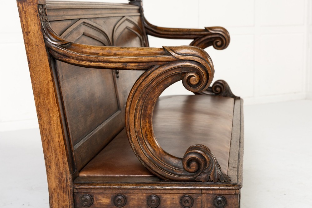 A nineteenth-century carved dark oak Gothic Revival bench featuring pointed arches on the backrest, scrolled armrests, and a fitted leather seat. View VI