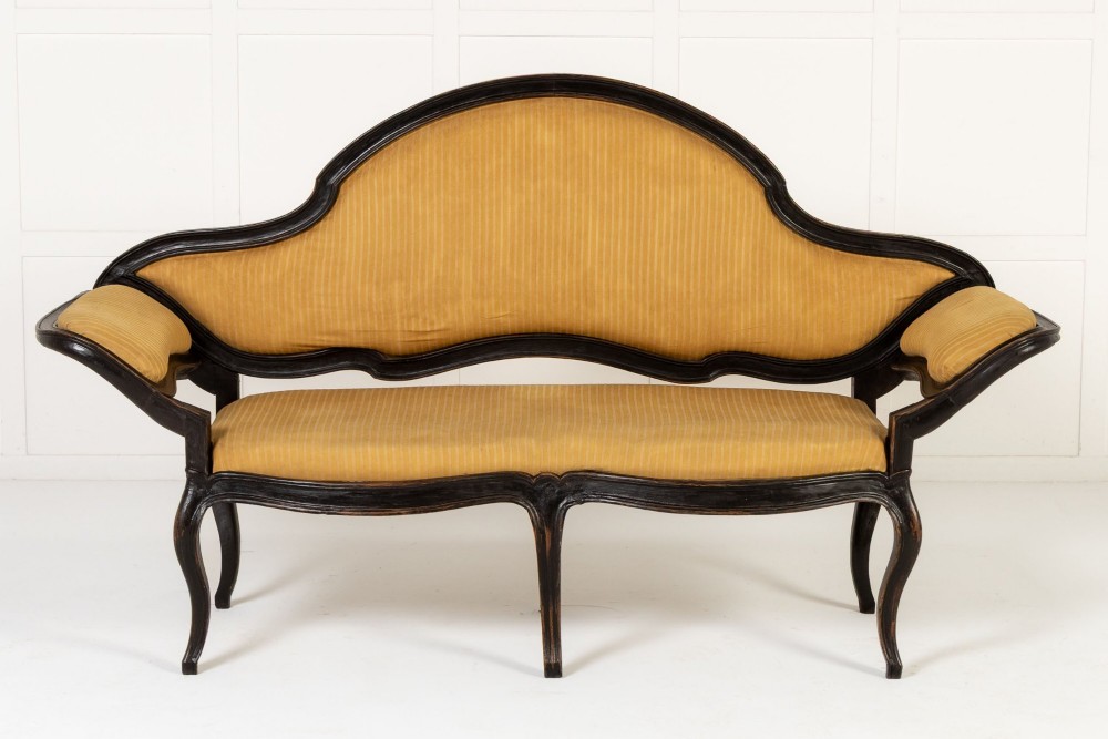 An 18th-century Italian ebonised sofa featuring a high serpentine backrest, mustard yellow striped upholstery, and carved cabriole legs.
