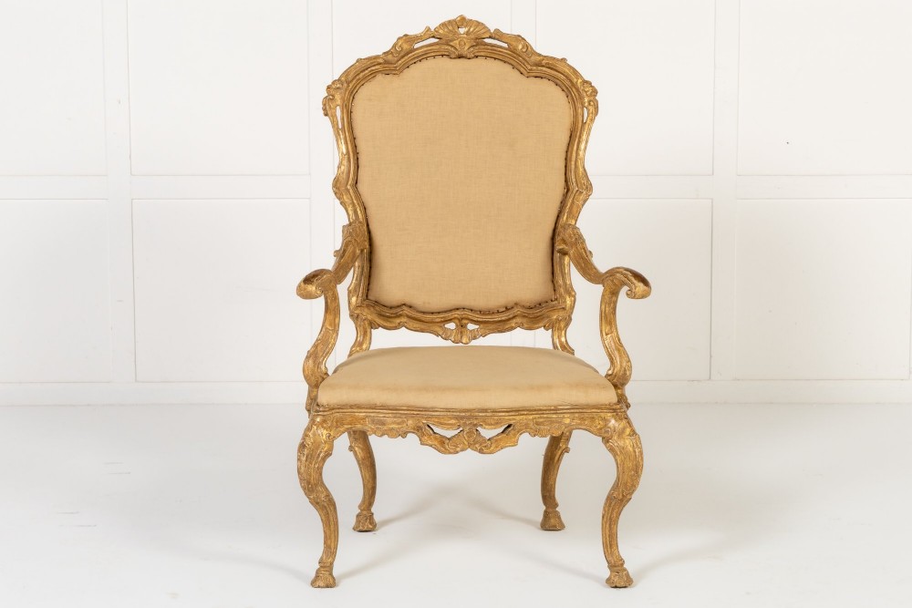 An 18th-century Italian carved giltwood armchair featuring neutral fabric upholstery, scrolled arms, and cabriole legs.
