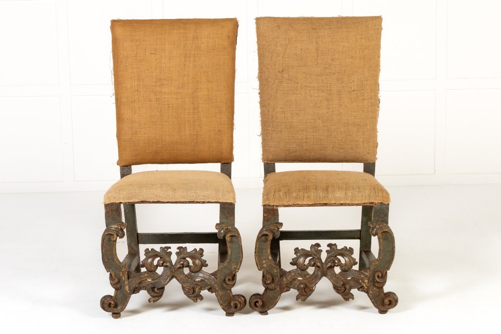 A pair of 17th-century Italian side chairs featuring tall backs upholstered in natural brown hessian, resting on elaborately carved scrollwork legs with a green painted finish.