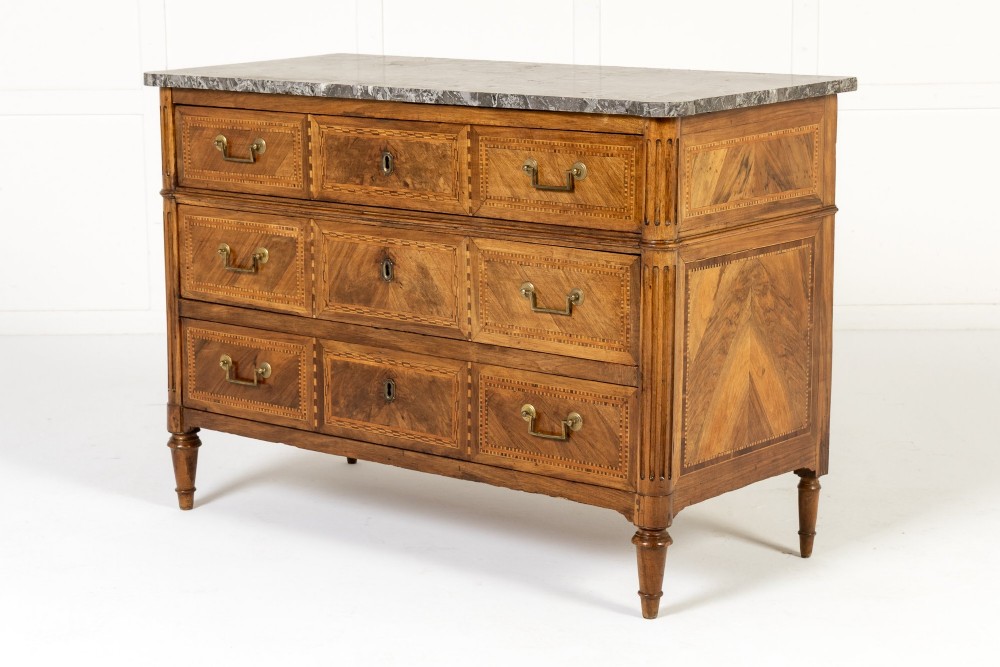 An 18th-century French walnut commode featuring three inlaid drawers, fluted corner pilasters, tapered legs, brass handles, and a grey marble top.