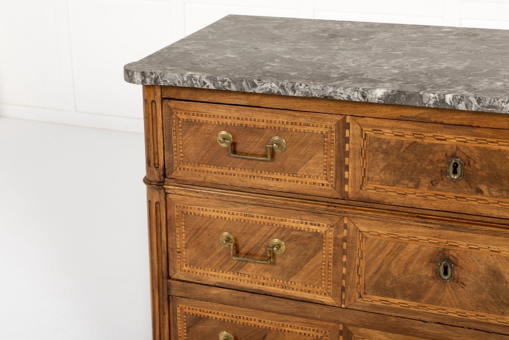 An 18th-century French walnut commode featuring three inlaid drawers, fluted corner pilasters, tapered legs, brass handles, and a grey marble top. View II