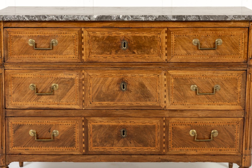 An 18th-century French walnut commode featuring three inlaid drawers, fluted corner pilasters, tapered legs, brass handles, and a grey marble top. View IV