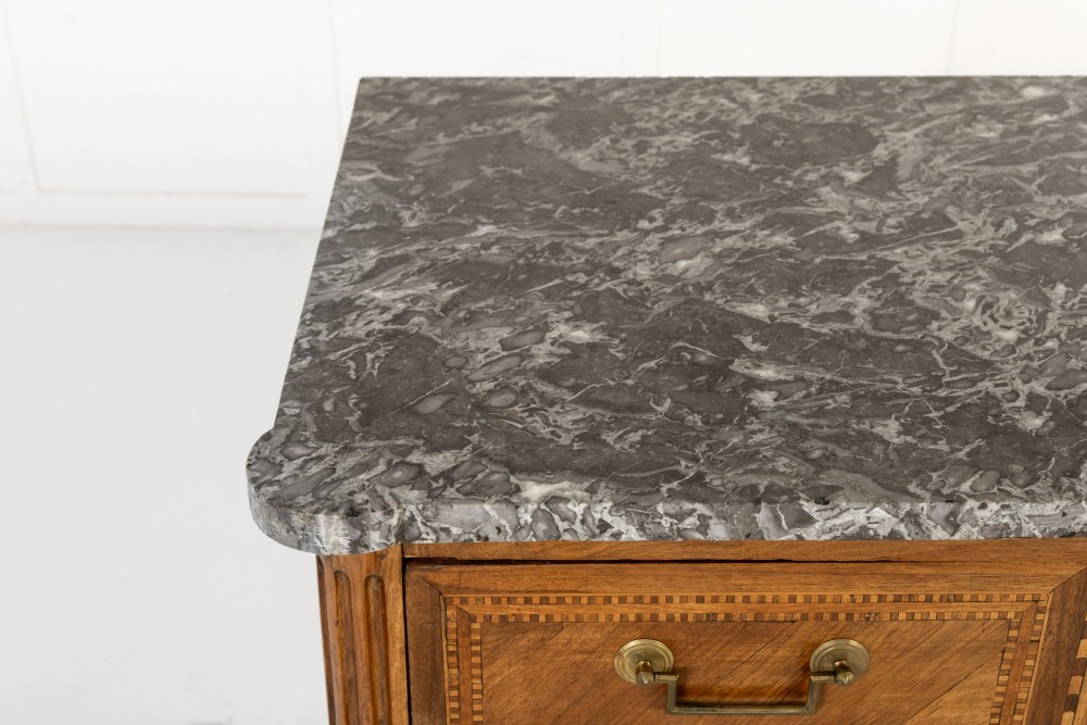 An 18th-century French walnut commode featuring three inlaid drawers, fluted corner pilasters, tapered legs, brass handles, and a grey marble top. View VII