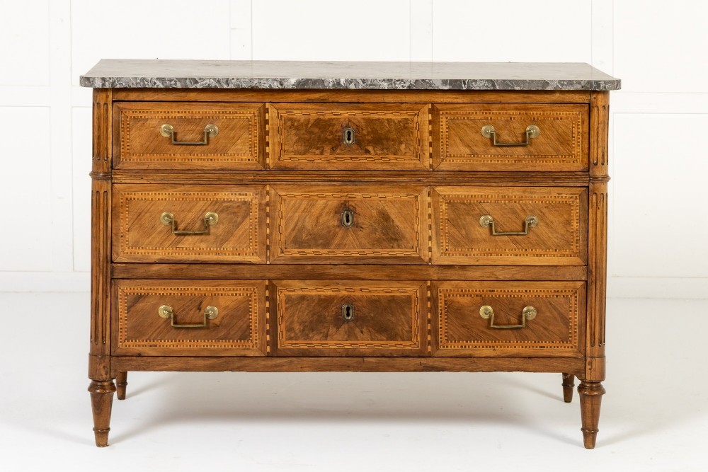 An 18th-century French walnut commode featuring three inlaid drawers, fluted corner pilasters, tapered legs, brass handles, and a grey marble top. View VIII
