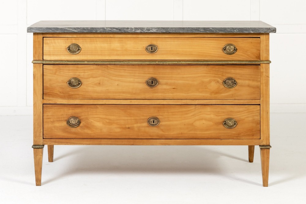 An 18th-century French cherrywood commode featuring three drawers with round brass ring pull handles, an oval keyhole escutcheon, tapered legs, and a grey marble top.