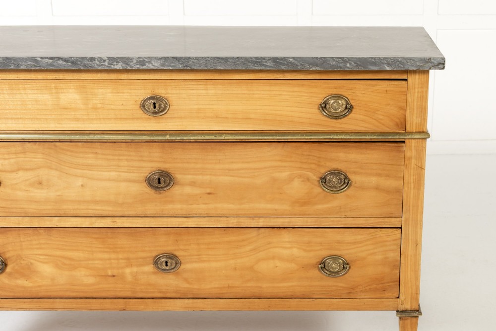 An 18th-century French cherrywood commode featuring three drawers with round brass ring pull handles, an oval keyhole escutcheon, tapered legs, and a grey marble top. View IV