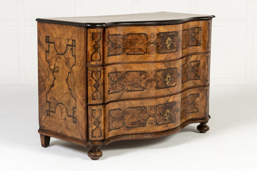 An 18th-century German walnut commode featuring three serpentine drawers with dark wood strapwork inlay and ornate brass escutcheons.