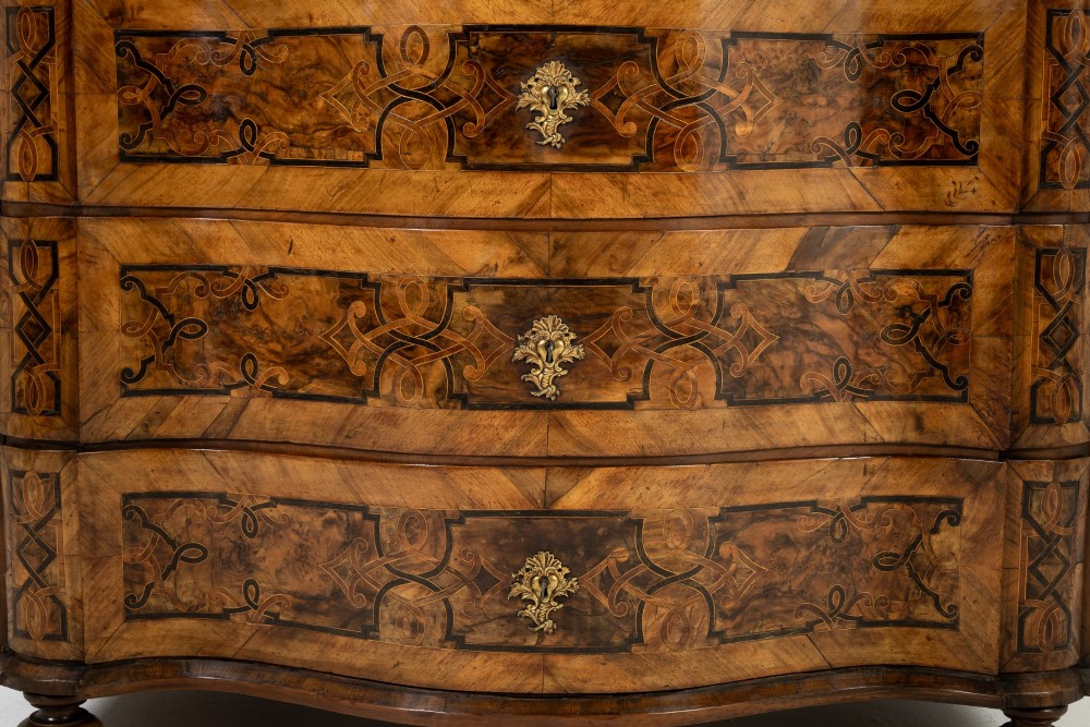 An 18th-century German walnut commode featuring three serpentine drawers with dark wood strapwork inlay and ornate brass escutcheons. View IV