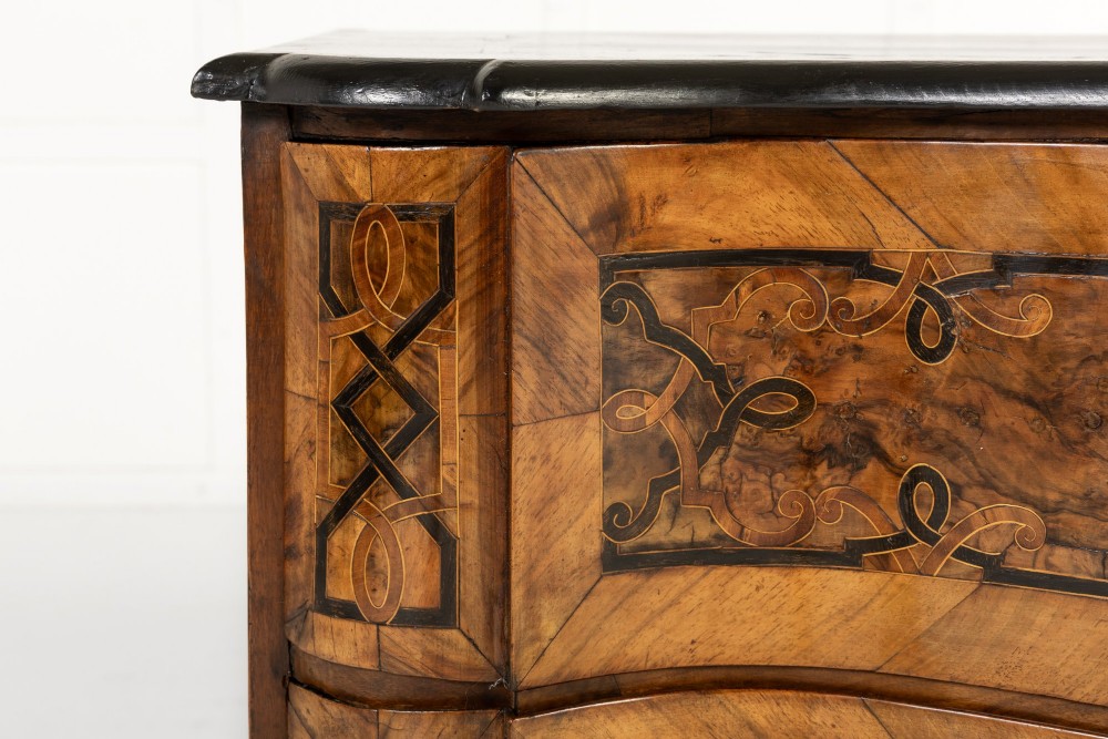 An 18th-century German walnut commode featuring three serpentine drawers with dark wood strapwork inlay and ornate brass escutcheons. View V