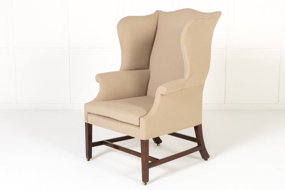 An 18th-century mahogany wingback armchair upholstered in neutral beige fabric with square timber legs and brass castors.