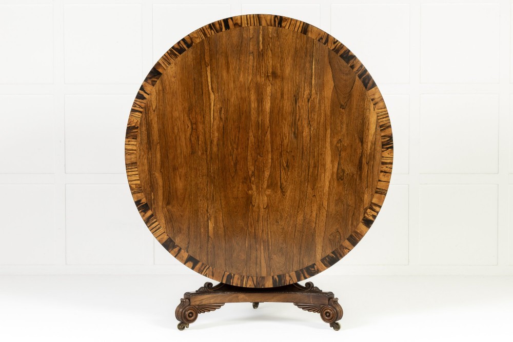A 19th-century English Regency circular rosewood centre table displayed with its crossbanded tilt-top tilted upright on a carved pedestal base with brass castors.