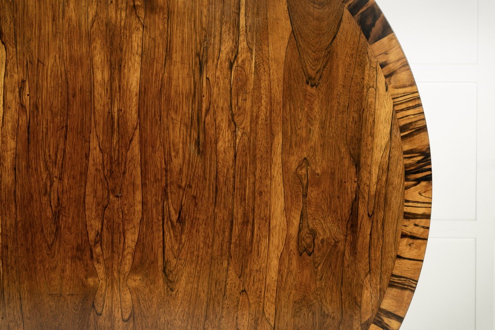 A 19th-century English Regency circular rosewood centre table displayed with its crossbanded tilt-top tilted upright on a carved pedestal base with brass castors. View II