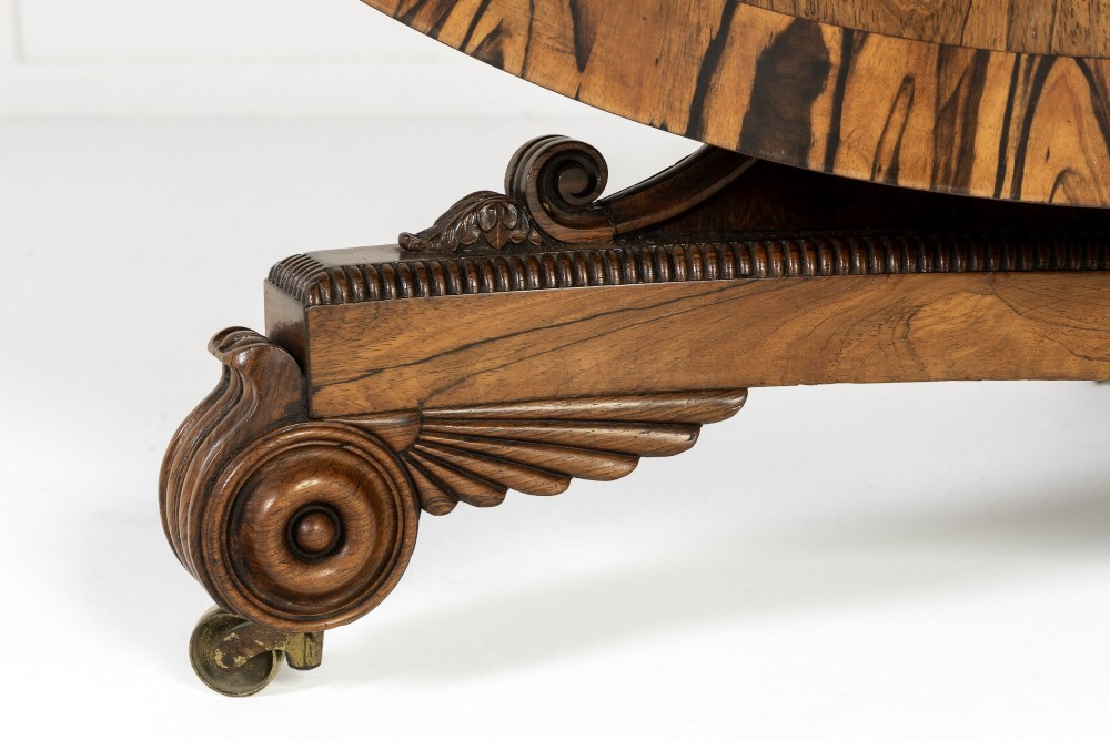A 19th-century English Regency circular rosewood centre table displayed with its crossbanded tilt-top tilted upright on a carved pedestal base with brass castors. View III