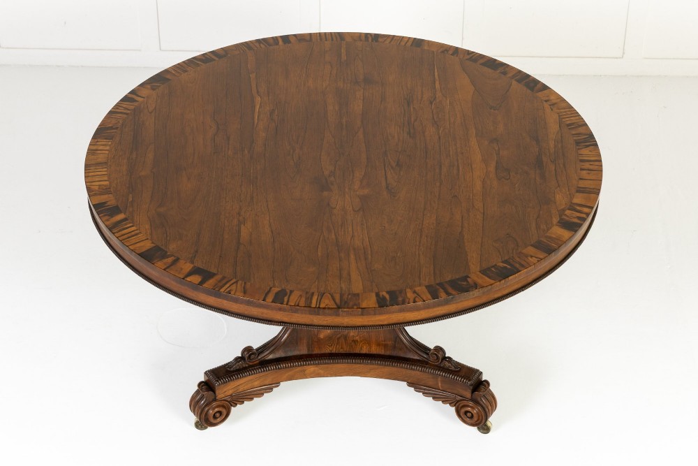 A 19th-century English Regency circular rosewood centre table displayed with its crossbanded tilt-top tilted upright on a carved pedestal base with brass castors. View IV