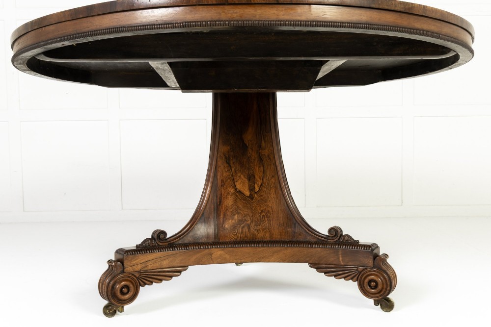 A 19th-century English Regency circular rosewood centre table displayed with its crossbanded tilt-top tilted upright on a carved pedestal base with brass castors. View V