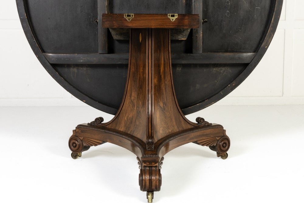 A 19th-century English Regency circular rosewood centre table displayed with its crossbanded tilt-top tilted upright on a carved pedestal base with brass castors. View VI
