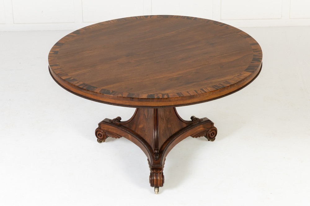 A 19th-century English Regency circular rosewood centre table displayed with its crossbanded tilt-top tilted upright on a carved pedestal base with brass castors. View VII