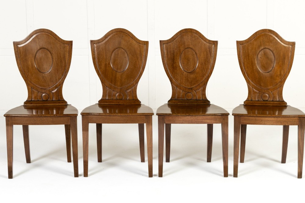 A set of four 19th-century Regency mahogany hall chairs featuring shield-shaped backs with carved oval medallions, solid timber seats, and square tapered legs.