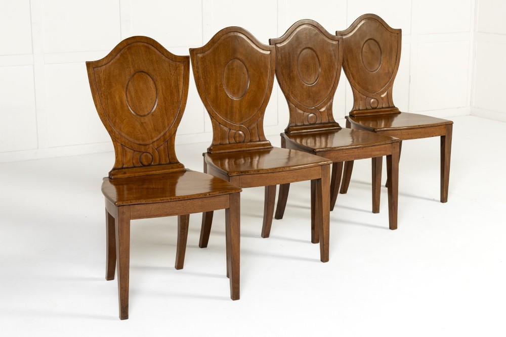 A set of four 19th-century Regency mahogany hall chairs featuring shield-shaped backs with carved oval medallions, solid timber seats, and square tapered legs. View II