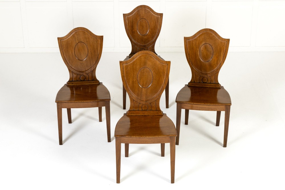 A set of four 19th-century Regency mahogany hall chairs featuring shield-shaped backs with carved oval medallions, solid timber seats, and square tapered legs. View III
