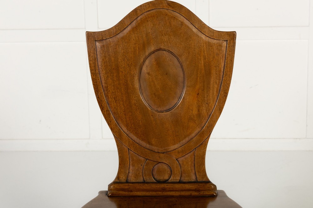 A set of four 19th-century Regency mahogany hall chairs featuring shield-shaped backs with carved oval medallions, solid timber seats, and square tapered legs. View VI