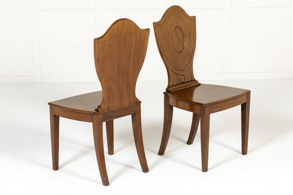 A set of four 19th-century Regency mahogany hall chairs featuring shield-shaped backs with carved oval medallions, solid timber seats, and square tapered legs. View IX