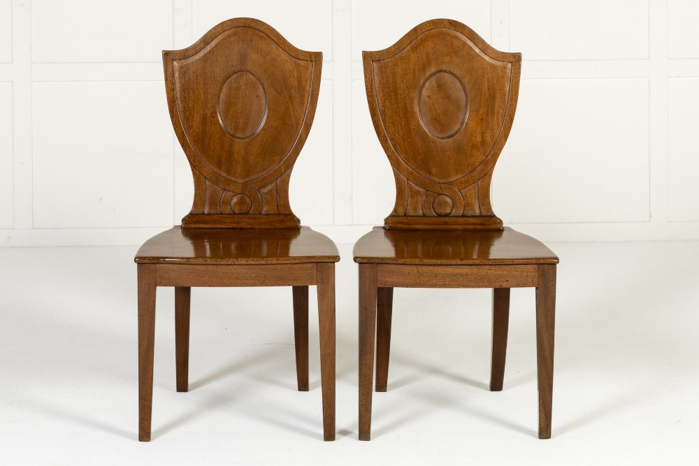 A pair of 19th-century Regency mahogany hall chairs with shield-shaped backrests, central oval cartouches, flat timber seats, and square tapered legs.