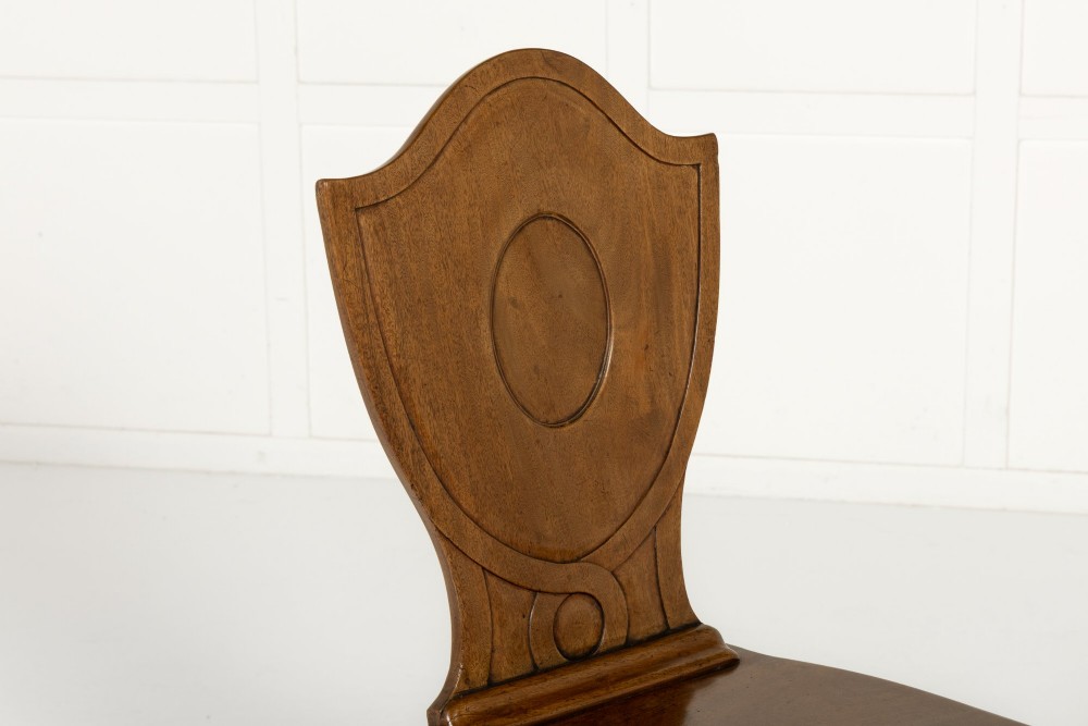 A pair of 19th-century Regency mahogany hall chairs with shield-shaped backrests, central oval cartouches, flat timber seats, and square tapered legs. View III