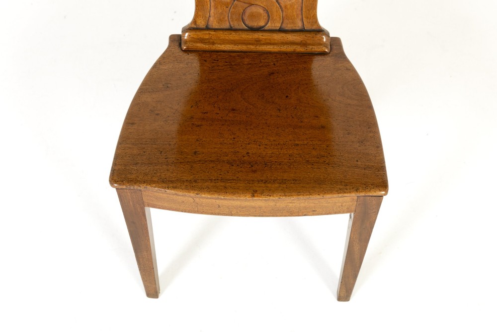 A pair of 19th-century Regency mahogany hall chairs with shield-shaped backrests, central oval cartouches, flat timber seats, and square tapered legs. View V