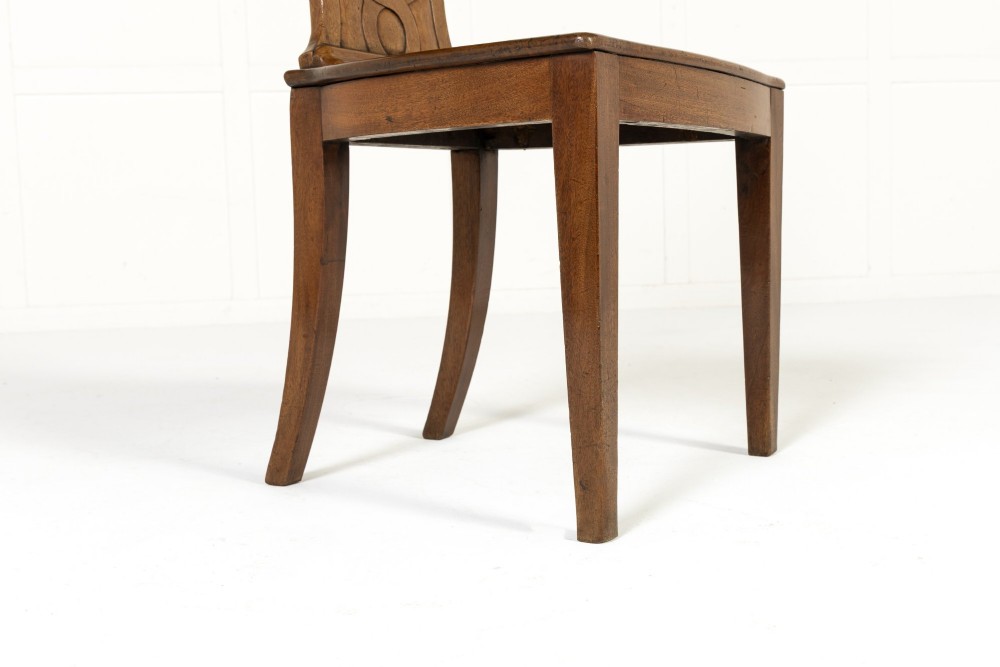 A pair of 19th-century Regency mahogany hall chairs with shield-shaped backrests, central oval cartouches, flat timber seats, and square tapered legs. View VI
