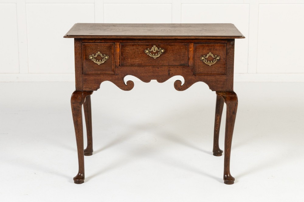 An 18th-century dark oak lowboy table featuring three drawers with brass handles, a shaped apron, and cabriole legs terminating in pad feet.