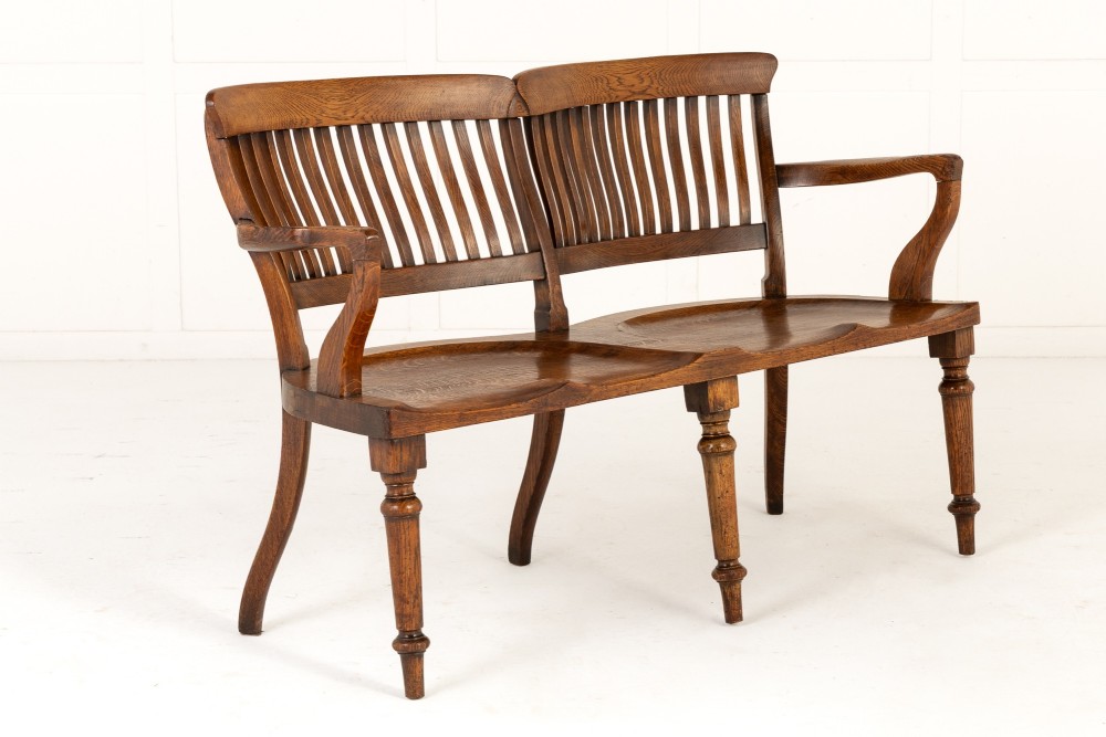 A 19th-century English oak double chair-back settee featuring twin slatted backrests, shaped seats, curved armrests, turned front legs, and splayed rear legs.