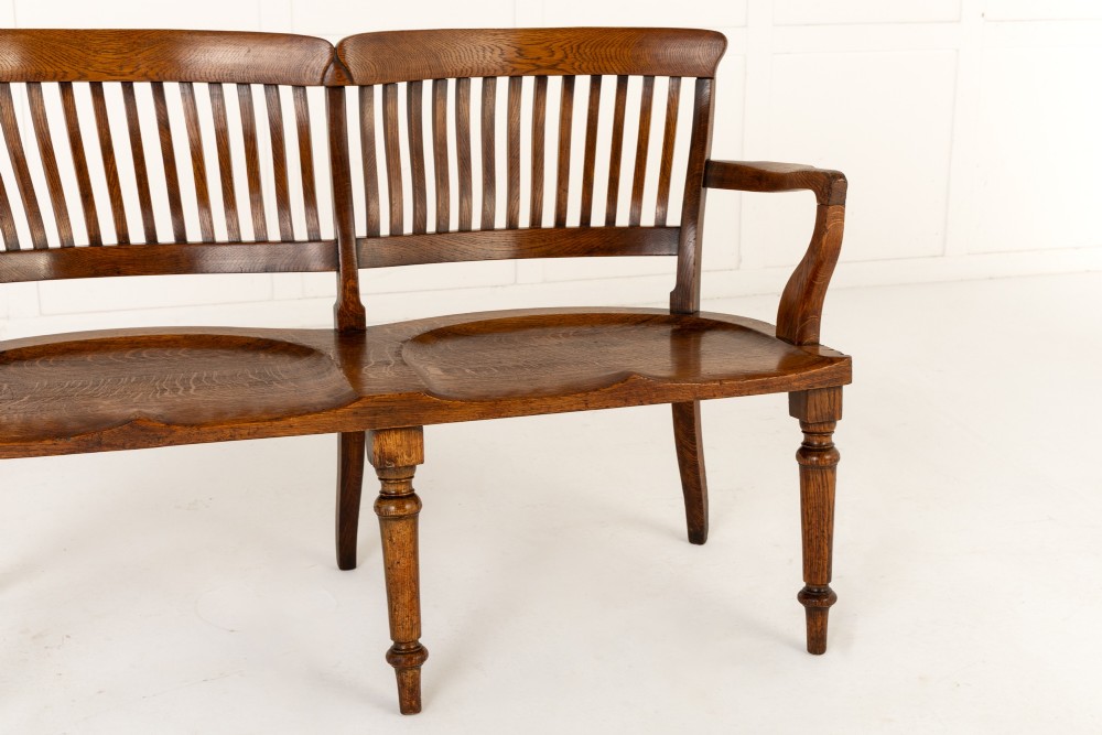 A 19th-century English oak double chair-back settee featuring twin slatted backrests, shaped seats, curved armrests, turned front legs, and splayed rear legs. View V