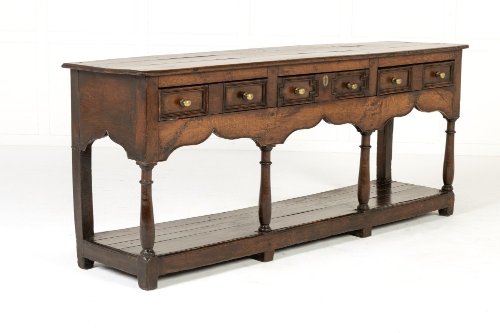 An 18th-century dark oak dresser base featuring geometric panelled drawer fronts, a shaped lower apron, turned front legs, and a bottom potboard shelf.