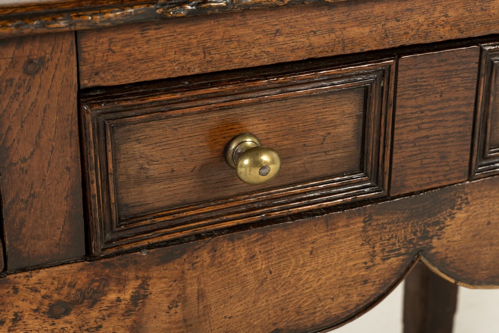 An 18th-century dark oak dresser base featuring geometric panelled drawer fronts, a shaped lower apron, turned front legs, and a bottom potboard shelf. View V