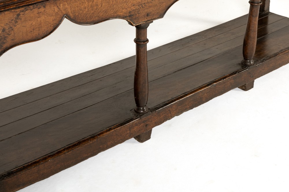 An 18th-century dark oak dresser base featuring geometric panelled drawer fronts, a shaped lower apron, turned front legs, and a bottom potboard shelf. View VI