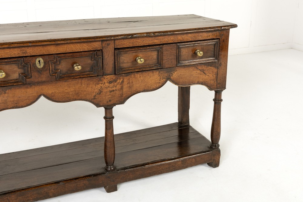 An 18th-century dark oak dresser base featuring geometric panelled drawer fronts, a shaped lower apron, turned front legs, and a bottom potboard shelf. View VII